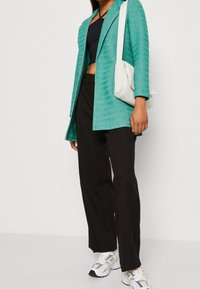 Türkiser strukturierter Blazer über einem schwarzen Croptop, kombiniert mit schwarzen weit geschnittenen Hosen und einer weißen Tasche. Die Turnschuhe haben graue und gelbe Akzente.