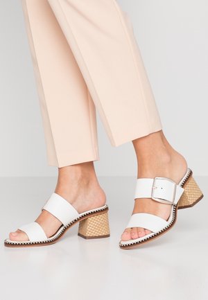 White heeled sandals with two wide straps, a large silver buckle, and a textured cork-style block heel. Smooth finish with decorative trim.