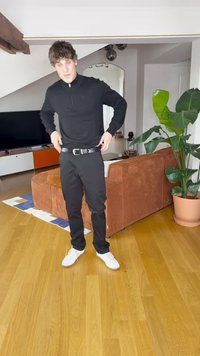 Sort genser med glidelås kombinert med svarte bukser, et svart belte med sølvspenne og hvite joggesko. Tregulv og en plante i bakgrunnen.