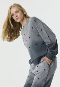 Grijze sweater en bijpassende sweatpants met zwarte stippen. De stof heeft een gladde textuur, met ribbelmanchetten en een roze logo-detail.