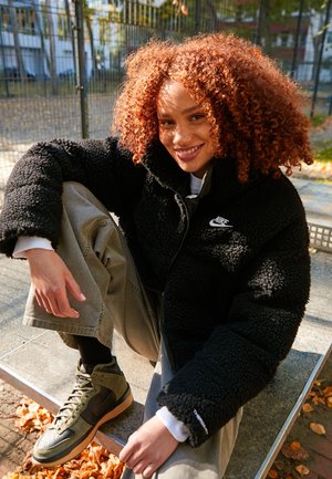 Femme souriante aux cheveux roux bouclés portant une veste en polaire noire Nike, un pantalon kaki et des baskets vertes, assise à l'extérieur sur une surface métallique.
