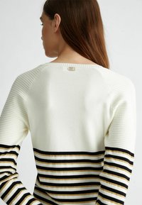 Maglione a righe con corpo bianco, texture a coste sulle spalle e righe orizzontali nere e dorate. Presenta un piccolo logo dorato sul retro.