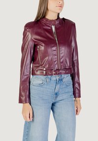 Veste courte en cuir bordeaux avec un col haut, fermeture éclair sur le devant, poches latérales et design ceinturé, portée sur un jean bleu clair.