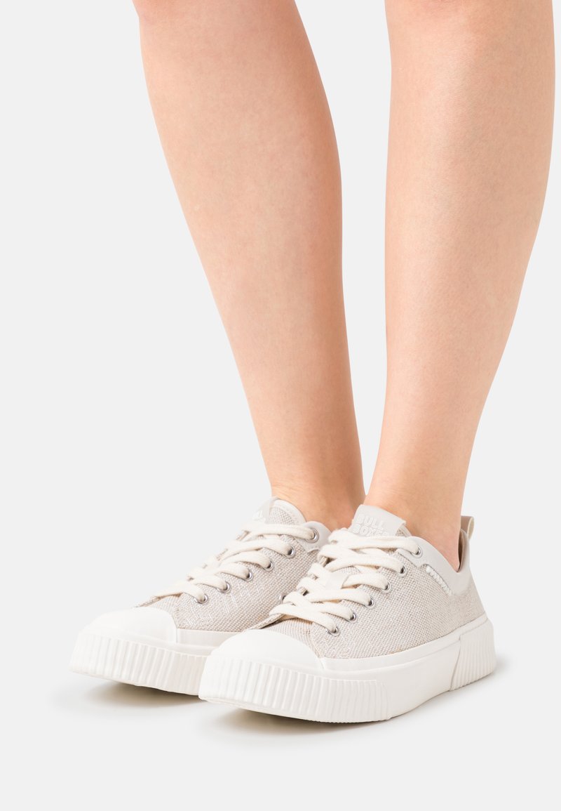 Lys grå kanvas sneakers med en teksturert overflate, hvit gummisåle og matchende snørebånd. Detalj av merkevaren logo på hæltappen.
