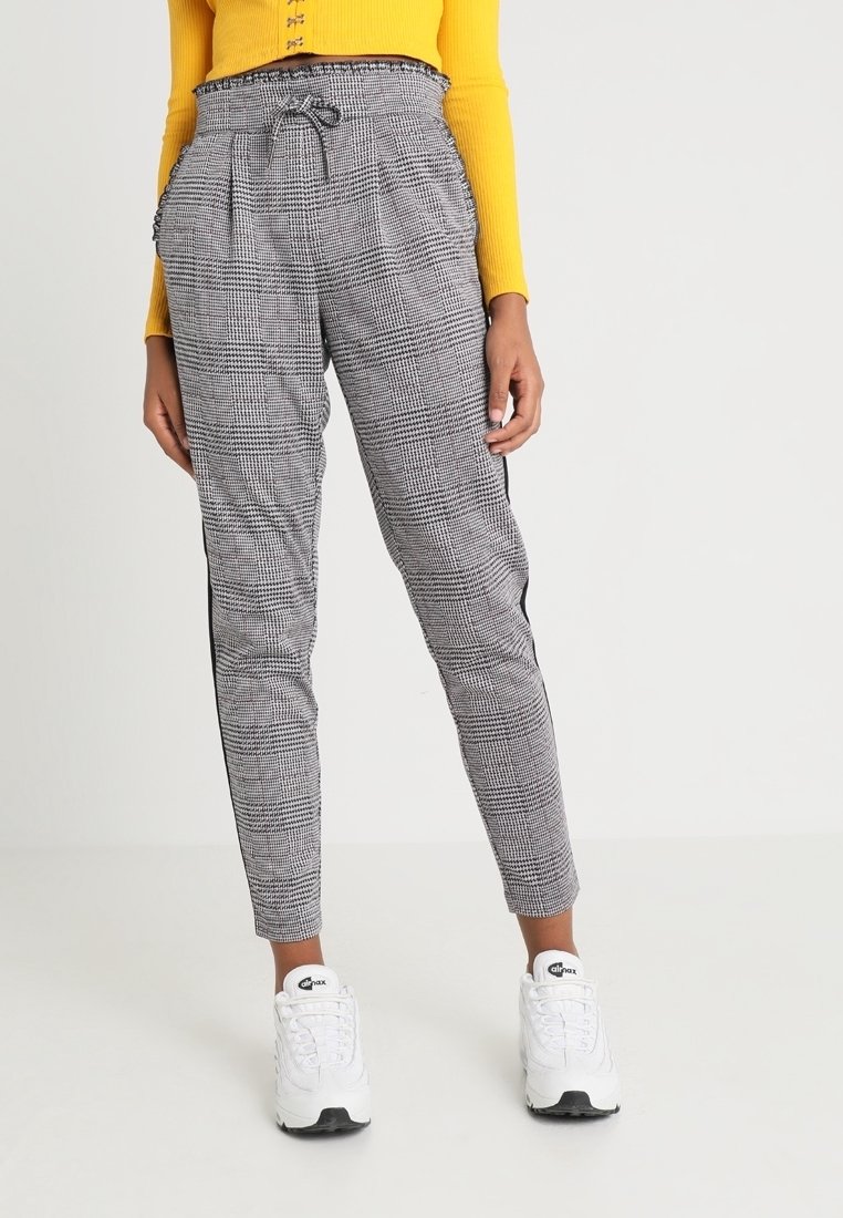 Femme portant un pantalon gris à carreaux effilés avec des rayures noires sur les côtés, un haut à manches longues jaune et des baskets blanches Nike Air Max.