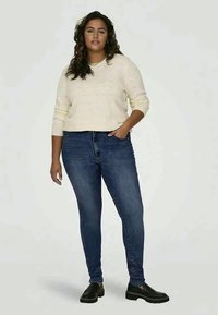 Krémy pletený sveter s textúrovaným vzorom, skombinovaný s tmavomodrými úzkymi džínsy a čiernymi koženými topánkami. Jednoduchý, neformálny outfit.