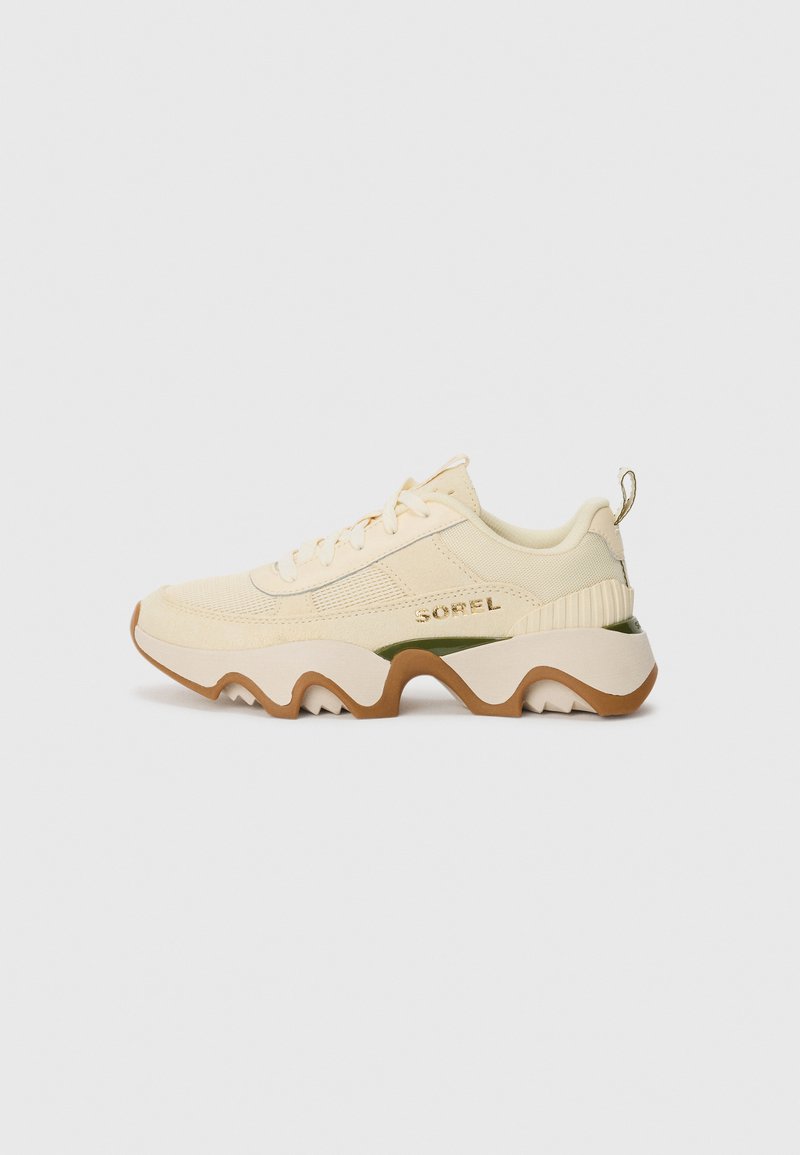 Kremfargede sneakers med en teksturert stoffoverdel, hvit og brun såle, og subtile grønne detaljer. Har rund tå og snøring.