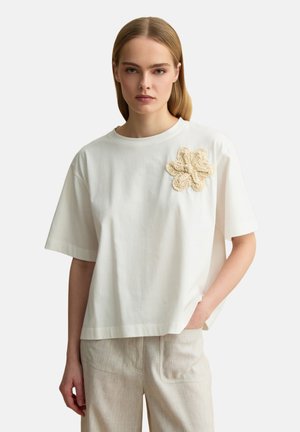 Vrouw met steil blond haar draagt een losse witte T-shirt met beige gehaakt bloemdetail en lichtbeige gestreepte broek, hand in zak.