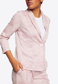 Světle růžový blazer s geometrickým vzorem, zapínání na jeden bílý knoflík a tříčtvrteční rukávy. Měkká látka s texturovaným povrchem.
