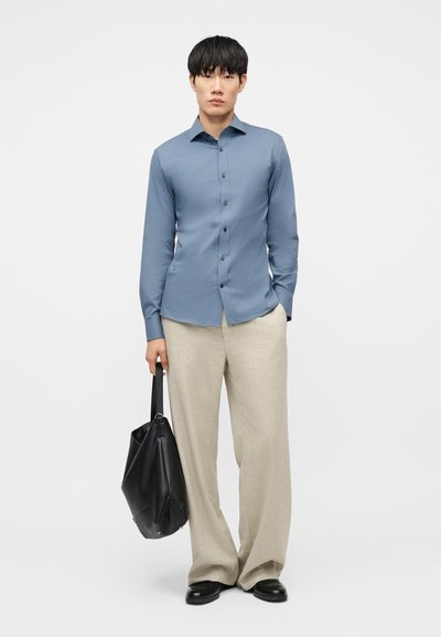 Νέος άνδρας που φοράει μπλε πουκάμισο με κουμπιά και μπεζ παντελόνι με φαρδιά πόδια, κρατώντας μια μαύρη δερμάτινη τσάντα ώμου μπροστά σε απλό φόντο.