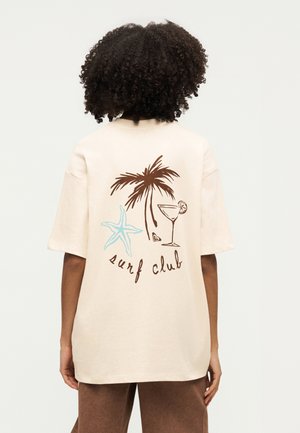 Osoba nosząca beżowy T-shirt z brązową palmą, niebieską rozgwiazdą, kieliszkiem koktajlowym i napisem „surf club” na plecach.