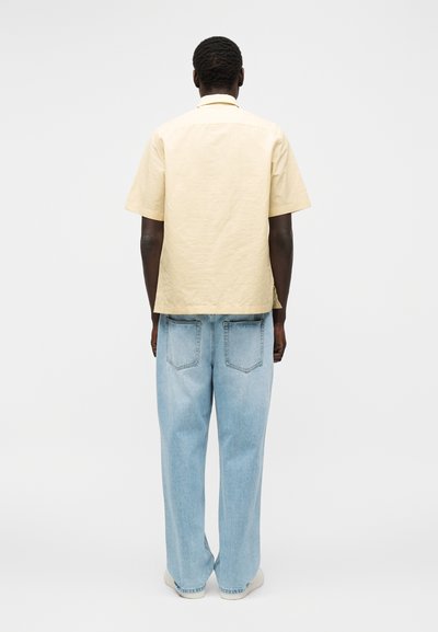 Άνδρας που στέκεται με την πλάτη, φοράει μπεζ κοντομάνικο πουκάμισο και ανοιχτό μπλε άνετο τζιν με λευκά παπούτσια σε μονόχρωμο φόντο.