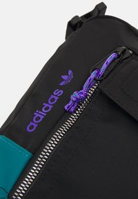 Svart tygryggsäck med en teal-akcent, lila Adidas-logotyp, en metallisk dragkedja och en flätad lila och blå snodd.