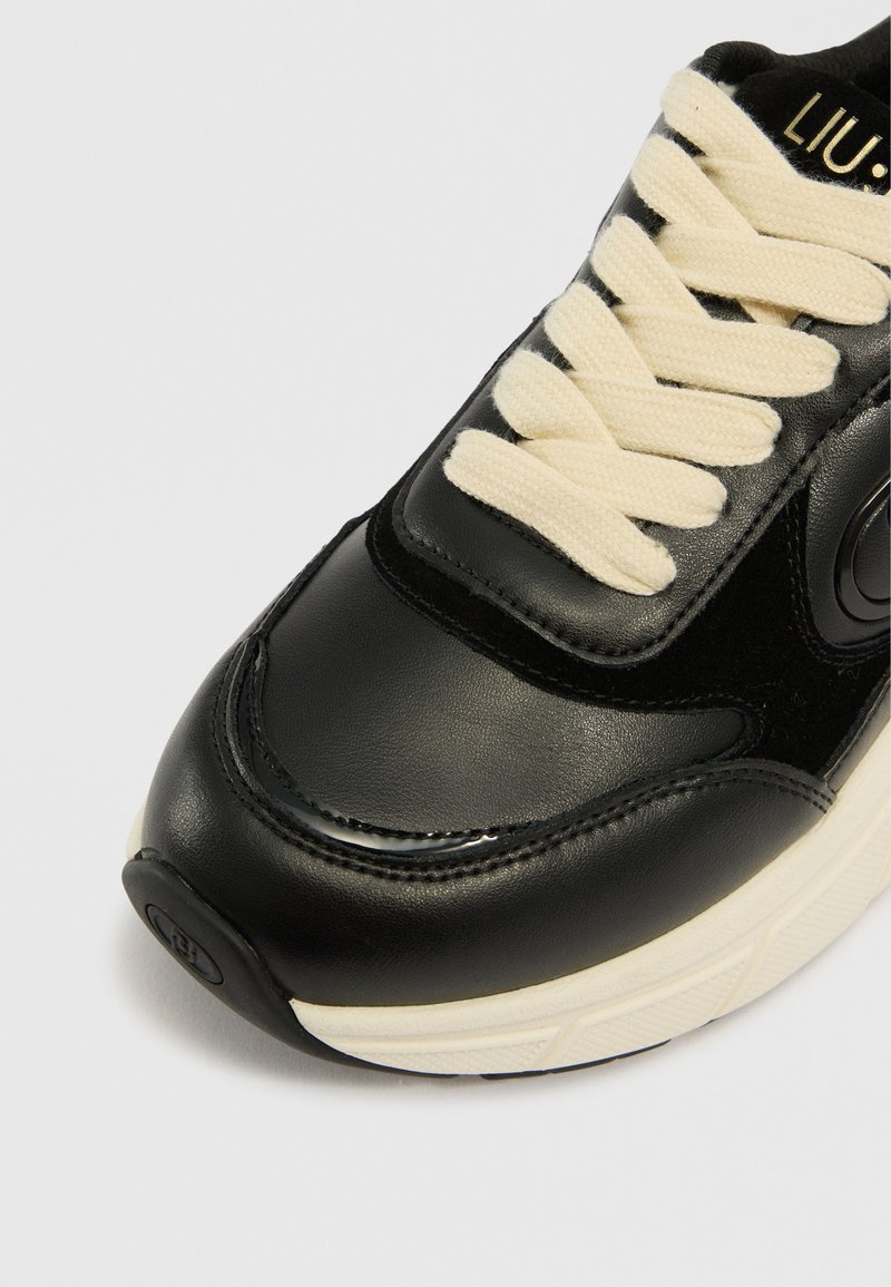 Schwarzer Sneaker mit einem glatten Obermaterial aus Leder und Wildleder, cremefarbenen Schnürsenkeln, runder Zehenpartie und strukturierten Sohle mit einem Logo an der Ferse.