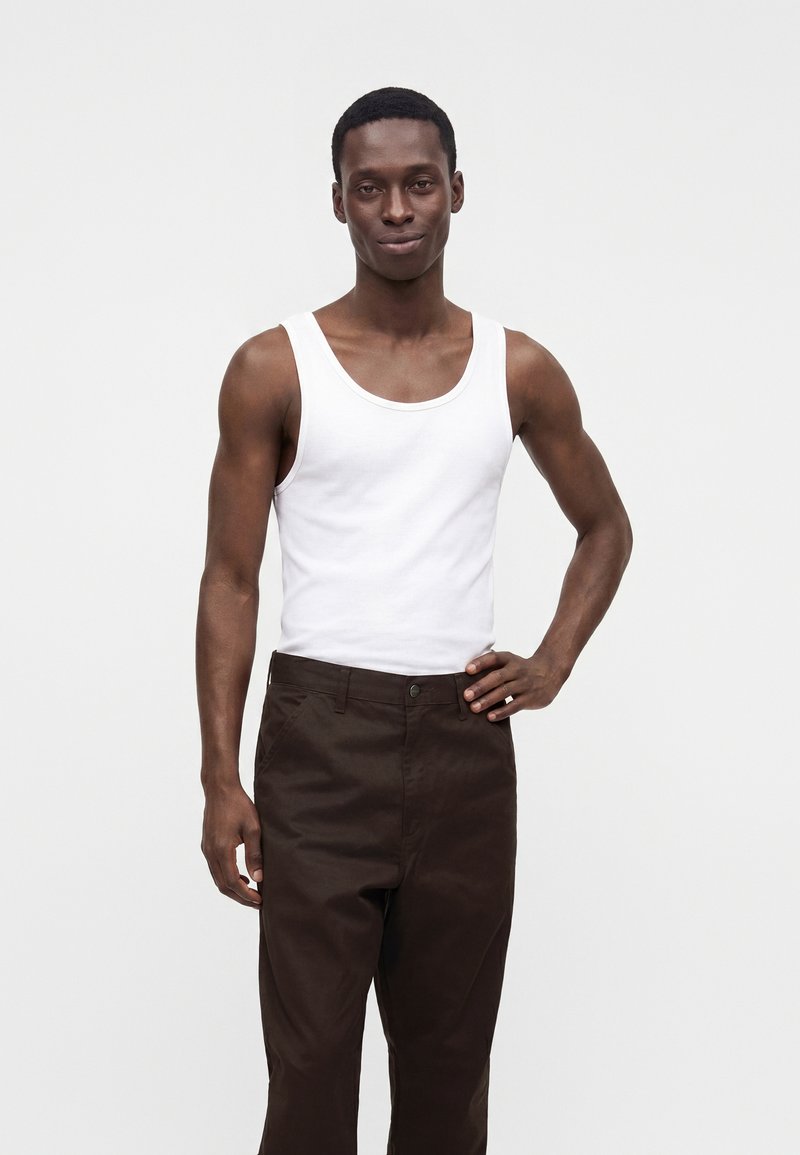 Jeune homme aux cheveux courts portant un débardeur blanc et un pantalon marron foncé, debout avec une main sur la hanche.