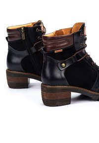 Botas de tobillo de cuero negro con acentos marrones, cremallera lateral, punta redonda y un tacón de madera texturizada. Detalle de cordones en la parte frontal.