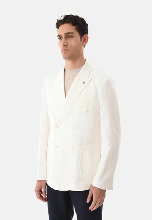 Uomo in piedi che indossa un blazer doppiopetto bianco sopra un maglione beige con pantaloni scuri su uno sfondo semplice.