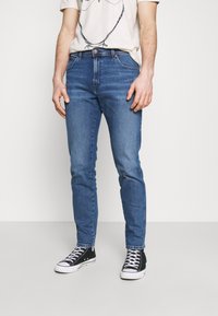 Blå denimjeans med slim fit, femficksdesign, kontrasterande sömmar och medelhög midja, parat med svarta sneakers.