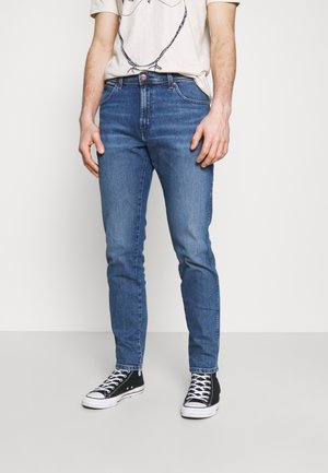 Blaue Jeans aus Denim mit schmaler Passform, Fünf-Taschen-Design, kontrastierenden Nähten und mittelhoher Taille, kombiniert mit schwarzen Sneakern.