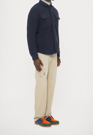 Mann trägt ein dunkelblaues Fleecehemd mit Knöpfen, beige Hose mit kleinen gestickten Mustern und mehrfarbige grüne, orange und blaue Schuhe.