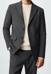 Blazer en laine gris avec revers crantés, deux boutons et une poche poitrine, assorti à un pull crème texturé et un pantalon gris assorti.