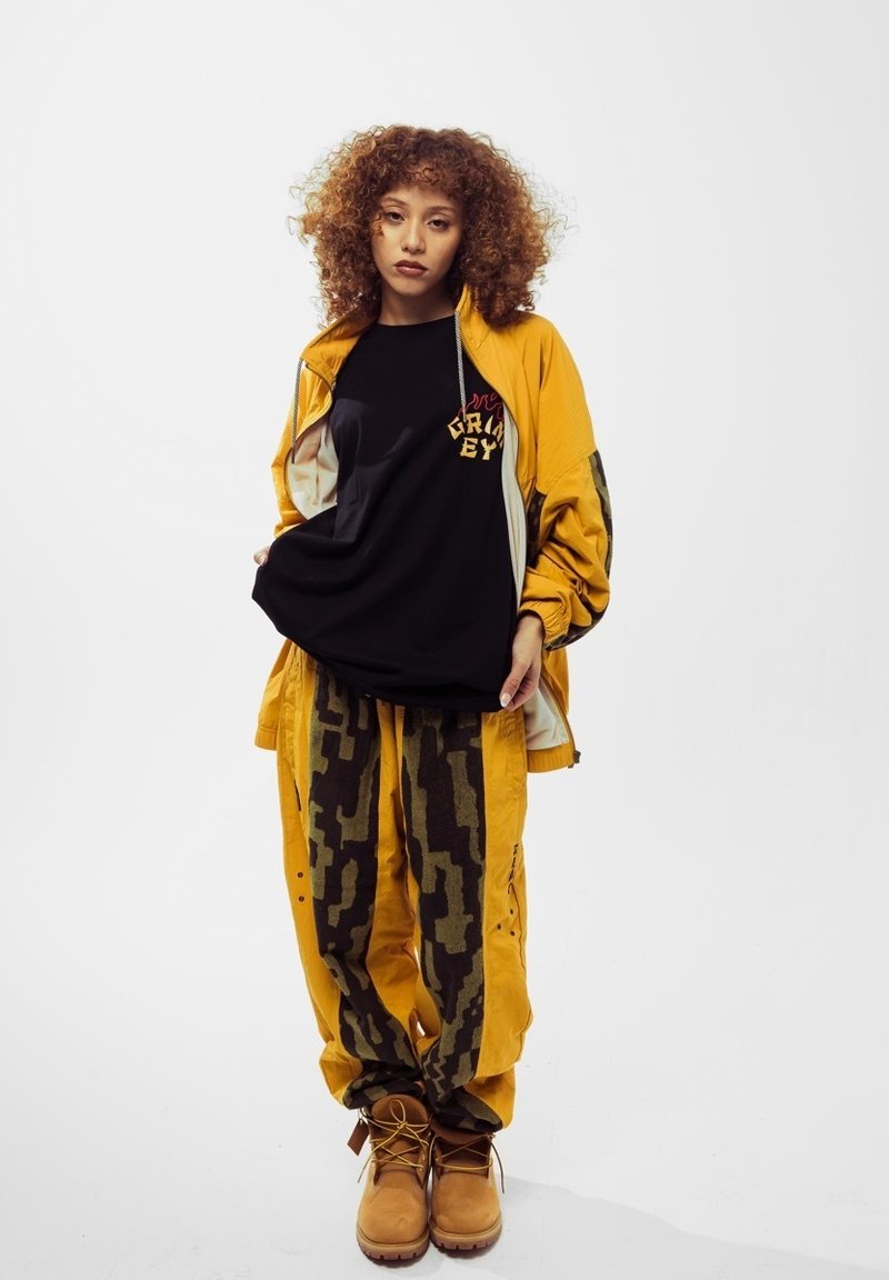 Yellow windbreaker with black accents, black T-shirt, and patterned yellow-brown camo pants. Brown boots complete the look.