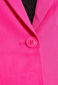 Růžový blazer s tvarovaným klopou, žebrovanou texturou a lesklým růžovým knoflíkem se čtyřmi otvory, který zdobí přední zapínání.