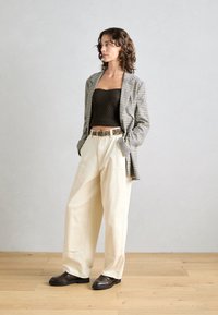 Blazer à carreaux, top côtelé marron, pantalon large crème et mocassins marron. Ceinture à imprimé léopard remarquée à la taille. Fond neutre.
