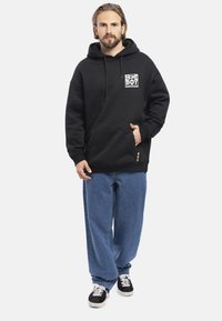 Felpa nera con tasca frontale, cappuccio con lacci e logo grafico. Abbinata a jeans blu larghi e scarpe nere con suola bianca.