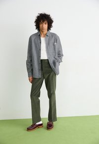 Geruite shirt in zwart en wit, groene corduroy broek, bordeauxrode schoenen met een brown zool, tegen een witte en groene achtergrond.