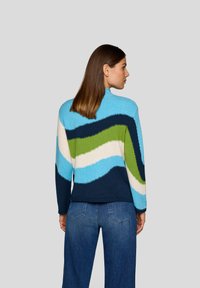 Frau mit glattem braunem Haar trägt einen bunt gestreiften Pullover in Blau, Grün, Weiß und Marine, kombiniert mit blauen Jeans, und steht seitlich.