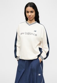 Γυναίκα με μακριά σκούρα μαλλιά, φοράει λευκό και ναυτικό μπλε φούτερ New Balance και ναυτικό μπλε αθλητικό παντελόνι, με τα χέρια στις τσέπες.