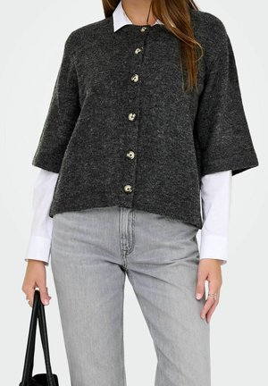 Femme portant un cardigan à manches courtes gris foncé sur une chemise blanche, jean gris clair, tenant un sac à main noir avec de longues anses.