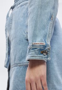 Μανίκι ανοιχτόχρωμου τζιν μπουφάν με ετικέτα Calvin Klein και λεπτομέρεια ασημένιου κουμπιού, που φοριέται από ένα άτομο με το χέρι του χαλαρά δίπλα στο σώμα.