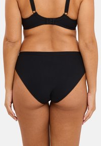 Slip bikini nero a coste con bordo ondulato, design a vita alta e copertura media sul retro. Texture morbida, fascia elastica in vita.