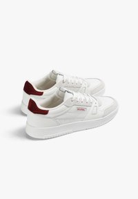 White low-top sneakers with red suede heel accents, perforated details, and branded "Scalpers" label on the side, displayed side by side.