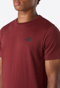 Nahaufnahme einer Person, die ein kastanienbraunes T-Shirt mit kurzen Ärmeln trägt, auf dessen Brust ein kleines schwarzes "The North Face"-Logo zu sehen ist.