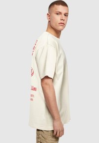 Beige cotton t-shirt with a relaxed fit, featuring red graphic text on the back and sleeve. Short sleeves and a round neckline.