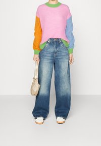 Person in lässigen, weit geschnittenen, blauen Jeans und einem farblich gemischten Pullover mit rosa, orange und blauen Ärmeln, hält eine beigefarbene Handtasche und trägt weiße Schuhe.