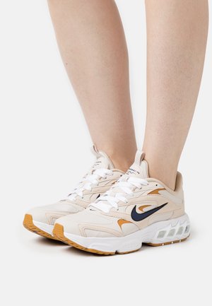 Beżowe sneakersy z fakturowaną cholewką, z czarnymi i pomarańczowymi akcentami, białymi sznurówkami, masywną podeszwą i widoczną jednostką poduszkową.