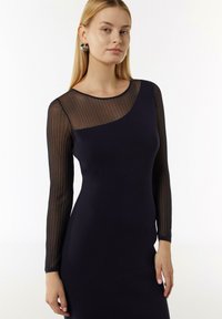 Granatowa sukienka o dopasowanej sylwetce i długich, przezroczystych, paskowanych rękawach. Posiada okrągły dekolt i gładką tkaninę. Elegancki i nowoczesny design.