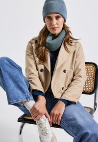 Giacca beige doppiopetto, berretto a coste blu, sciarpa coordinata, maglietta scura, jeans blu e sneakers bianche con accenti beige. Seduto su una sedia.