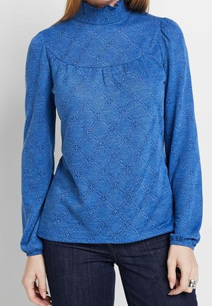 Vrouw draagt een blauwe blouse met lange mouwen, met geponste bloemmotieven en een geplooide hoge hals, gecombineerd met donkere spijkerbroek.
