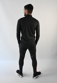 Camiseta negra deportiva de manga larga y pantalones ajustados con detalles en dorado. Los pantalones tienen cremalleras azules y rojas en los tobillos. Modelo de espaldas.