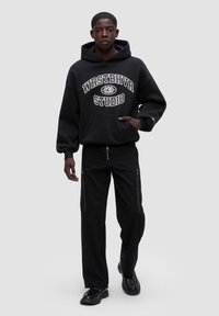 Černá mikina s kapucí a bílým nápisem "WESTHAVER STUDIO", v kombinaci s černými kalhotami se zipovými detaily. Hladká látka, uvolněný střih.