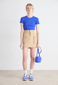 T-shirt aderente blu, gonna cargo beige con tasche, borsa blu, calze bianche, sneaker blu e bianche, sfondo neutro.