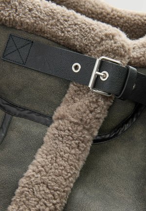 Nahaufnahme eines grauen Schaffellmantels mit weicher Wollfütterung und einem schwarzen Lederband, das mit einer silbernen Schnalle gesichert ist.