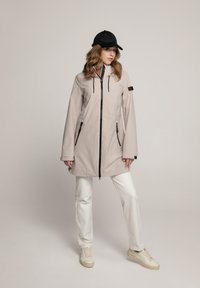 Lichtroze waterdichte jas met zwarte ritsen en accenten, twee voorzakken en een capuchon. Gelaagd met witte broek en schoenen.
