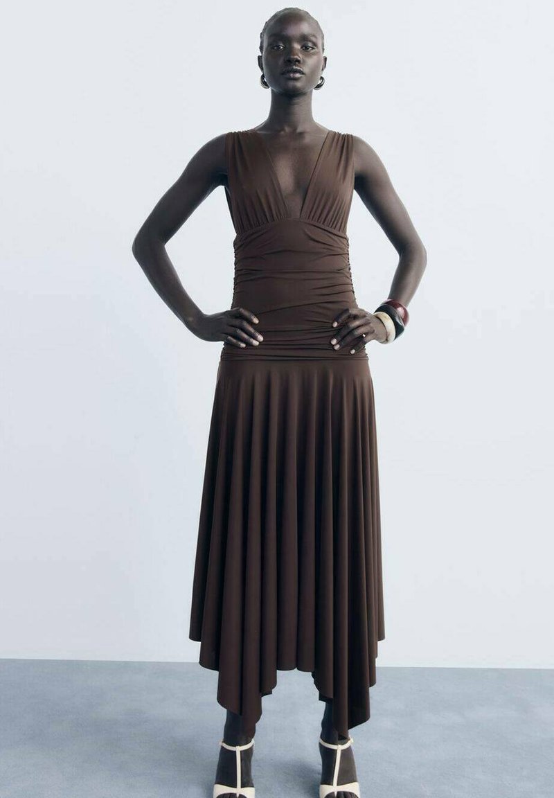 Robe marron sans manches avec un décolleté en V profond et un corsage froncé. La jupe présente des plis et un ourlet asymétrique. Le mannequin porte des bracelets imposants.