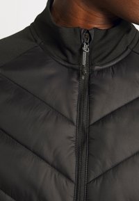 Gros plan d'une veste matelassée noire avec col haut et fermeture éclair, portée par une personne montrant une partie de son cou et de sa mâchoire.
